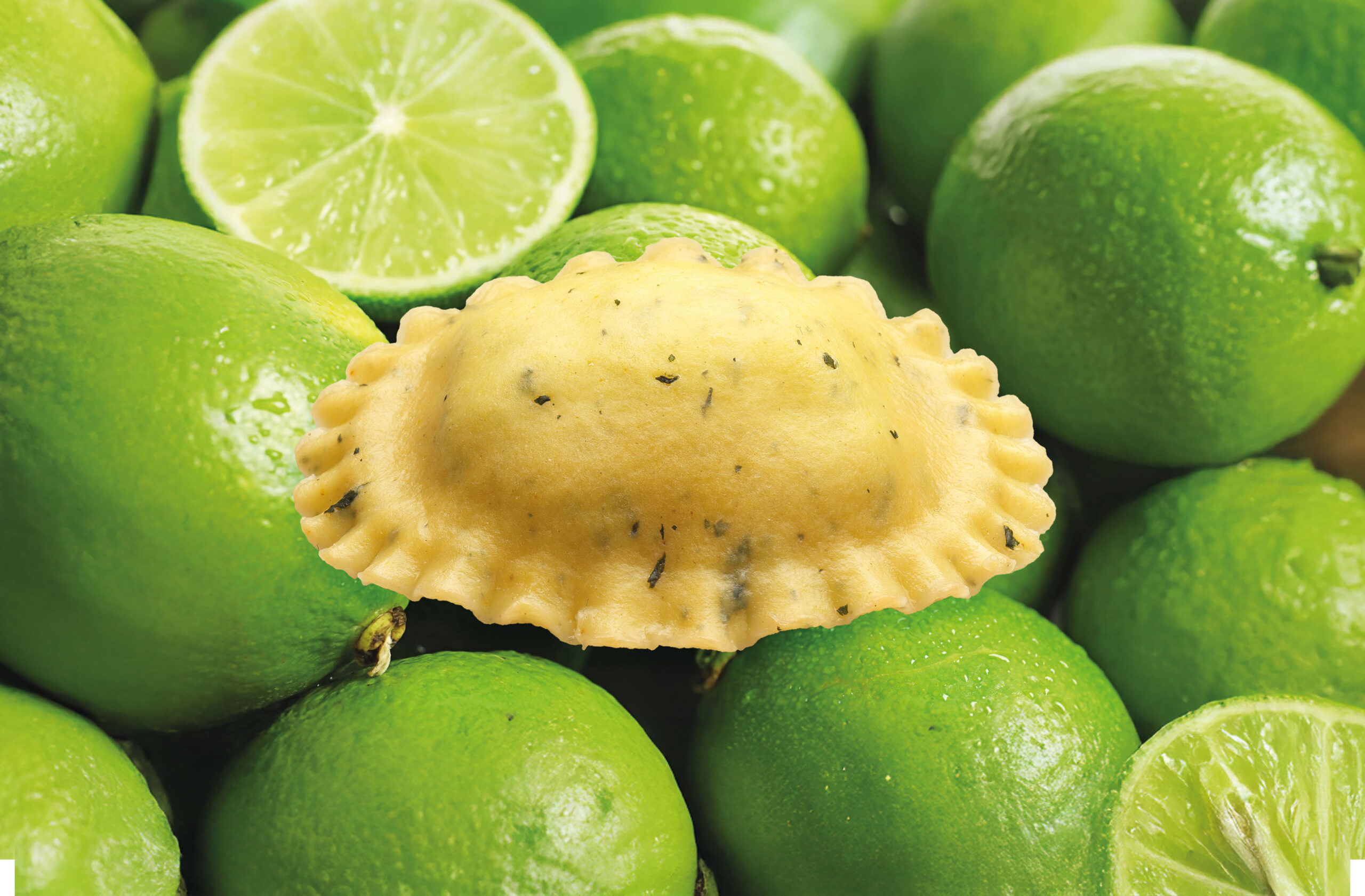 In der Mitte befindet sich ein einzelnes Stück Ravioli-Nudeln, umgeben von ganzen und in Scheiben geschnittenen grünen Limetten im Hintergrund. Die Ravioli sind scharf abgebildet, während die Limetten leicht verschwommen sind.