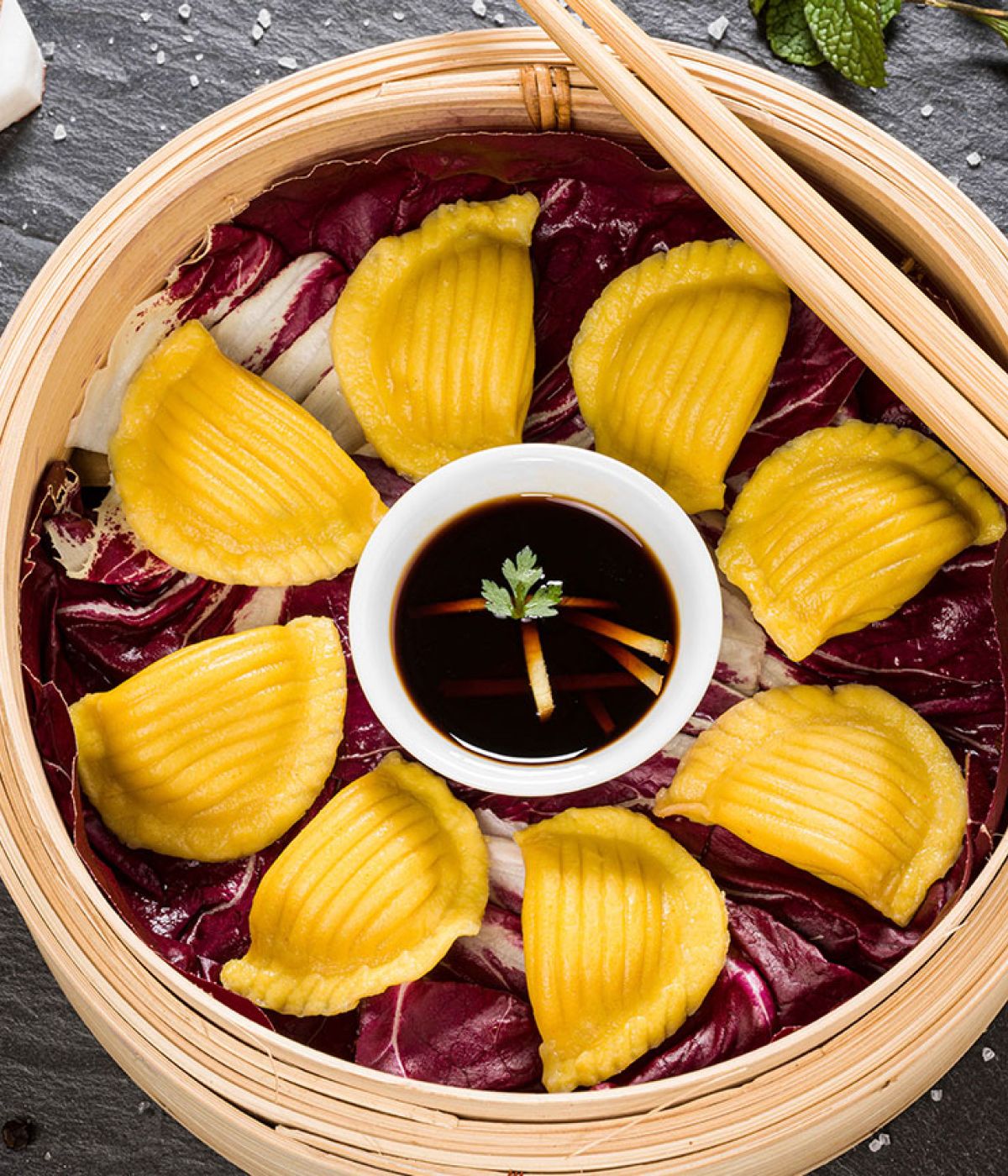 Ein Bambusdämpfer, gefüllt mit gelben Teigtaschen, die kreisförmig um eine Schale mit Sojasauce angeordnet sind.